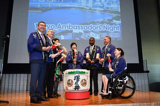 Governor Koike stands on stage flanked by six ambassadors